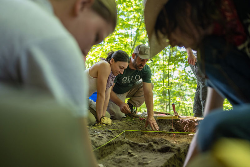 A professor works with students at an archeological dig site in the forest.