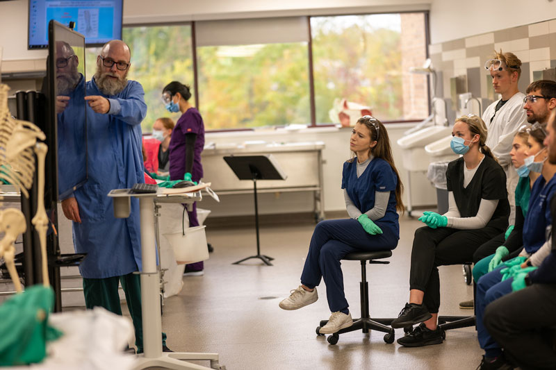 Medical students wearing scrubs listen during a lecture from a professor.