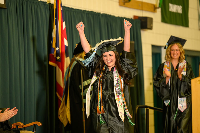 A student celebrates while walking across the stage during graduation, wearing her cap and gown.